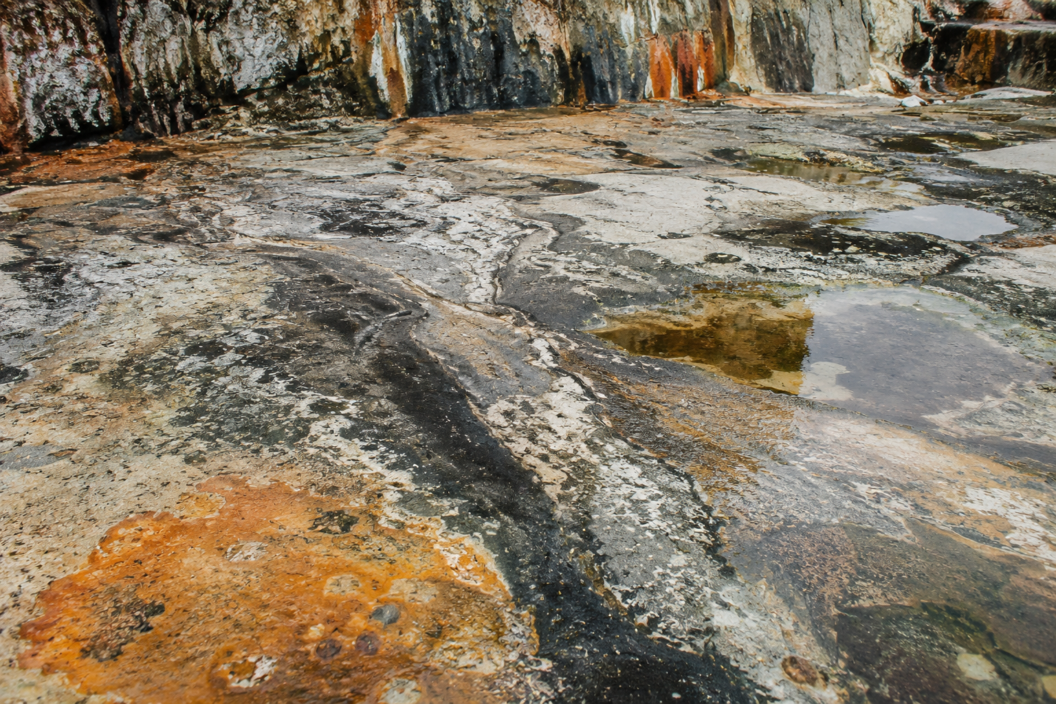 Eroded coastal rock and concrete showing cracks, mineral stains, salt marks and reflective pools.